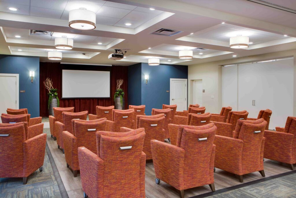 Sonata West 2018 - The Summit of Winter Garden A movie theater room with rows of orange sofa chairs facing the white projector screen at The Summit of Winter Garden, a senior living community near Clermont, FL.