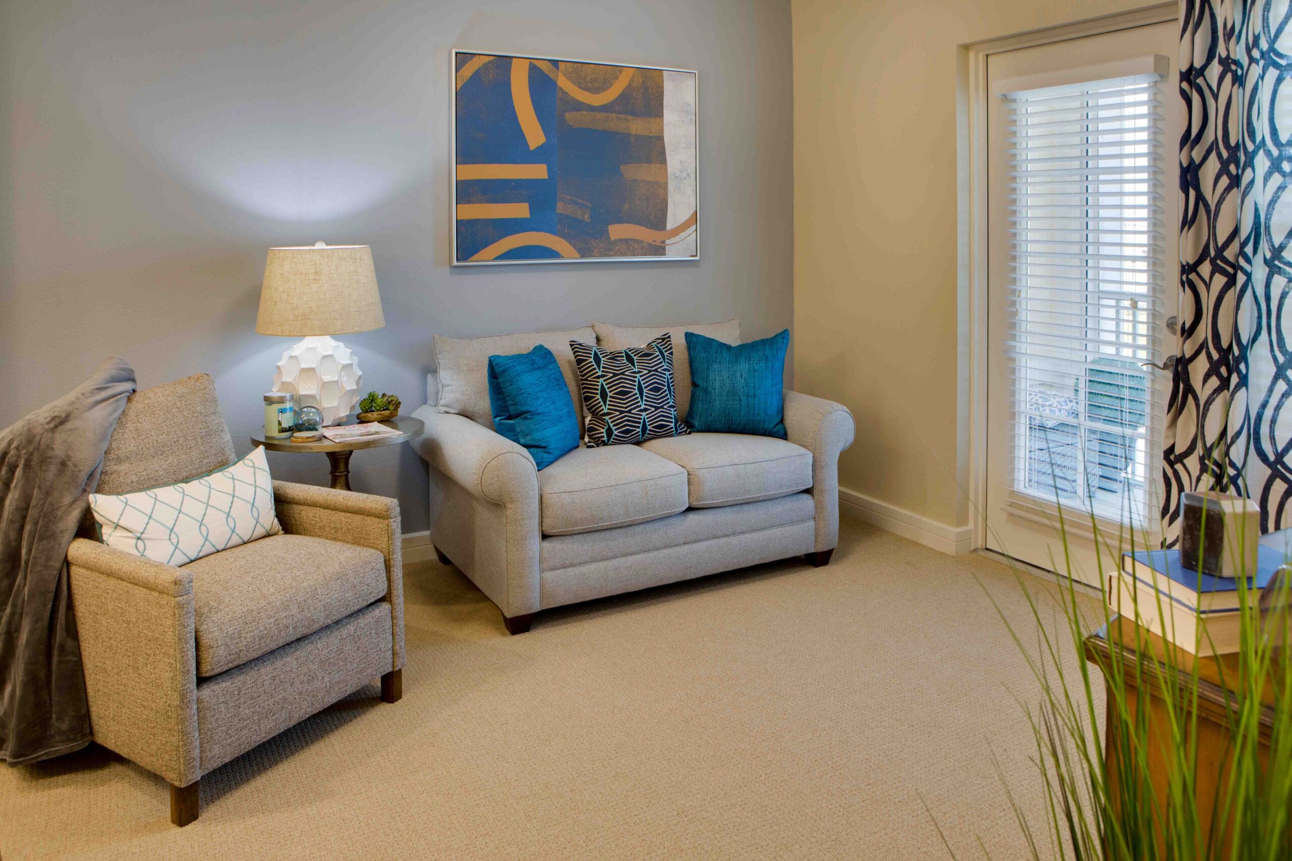 Sonata West 2036 - The Summit of Winter Garden A living room with a gray couch with blue throw pillows, a beige sofa chair, a side table with a lamp on it, and a window leading to the balcony with blue curtains at The Summit of Winter Garden, an independent and assisted living community near Orlando.