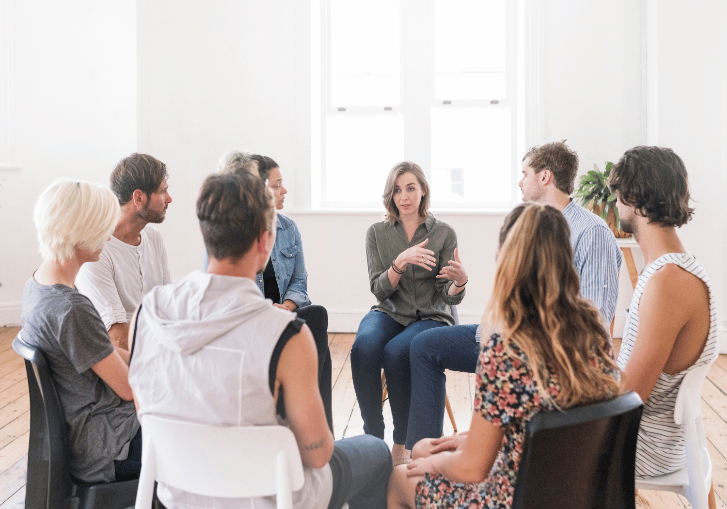 A woman leads a group discussion in a circle of seated adults in a bright room while others listen attentively.