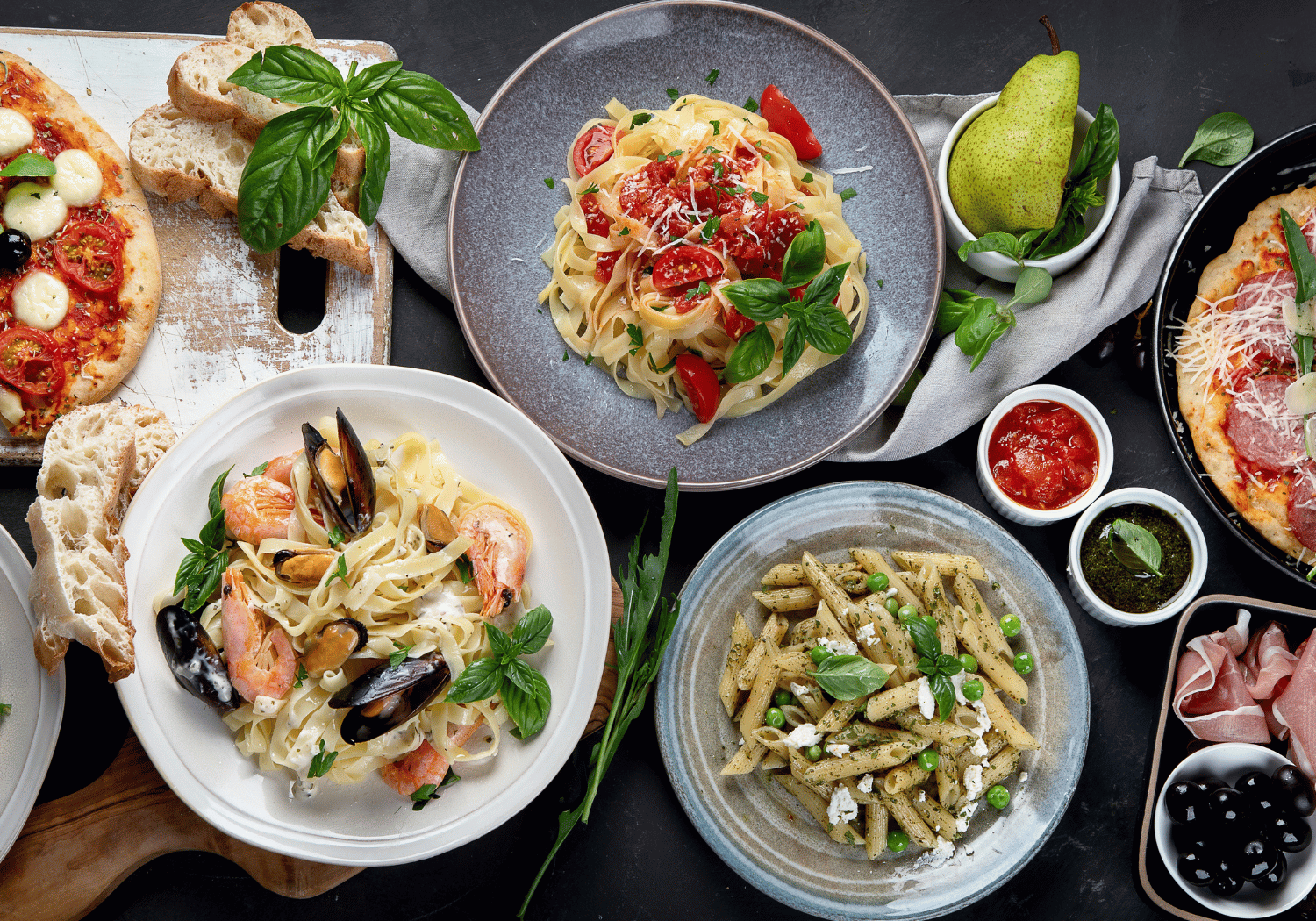 A colorful spread of Italian dishes on a dark table: seafood pasta, tomato pasta, pizza slices, crusty bread, and a pear.