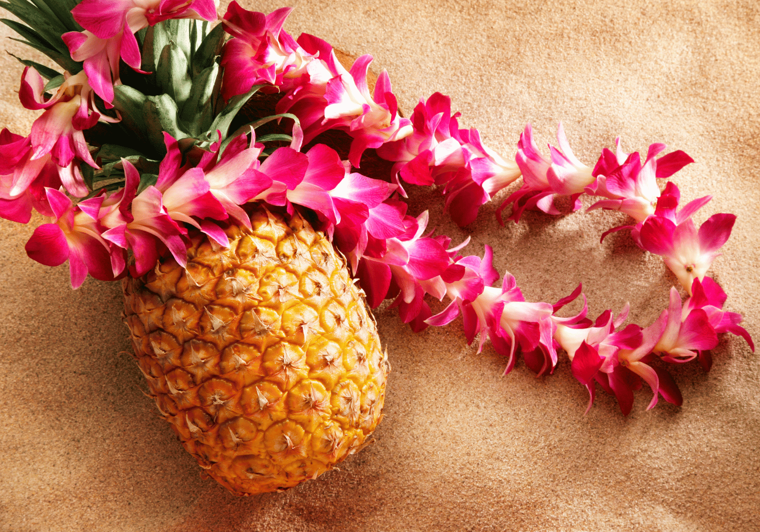 Pineapple with a pink flower lei on sandy beach surface.