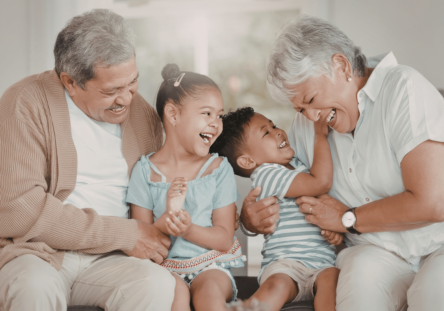 Grandparents laughing with two young grandchildren as they sit together on a couch in a bright room.